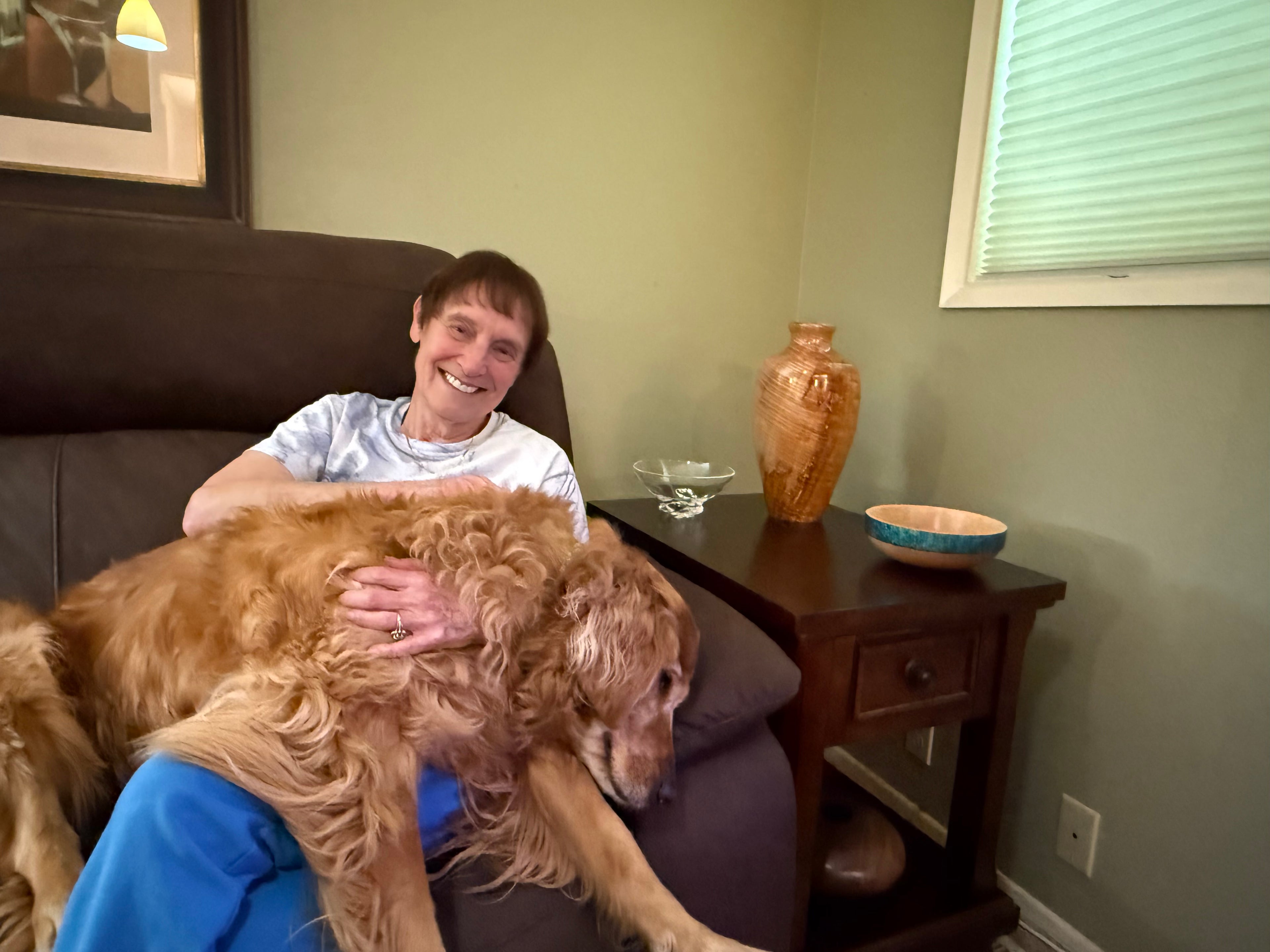 Person holding a large dog in a living room with a side table and decorative items.